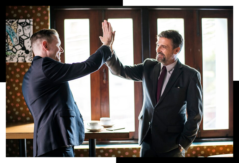 Group 7 Two professional men in business suits exchanging a high-five, symbolizing collaboration and success in a commercial setting, with coffee cups on a table in the background.