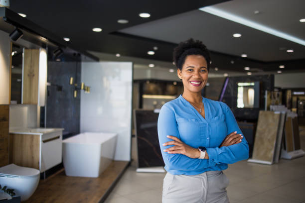 Smiling woman in a blue blouse standing confidently in a modern bathroom showroom, showcasing stylish fixtures and design options for affordable remodeling.