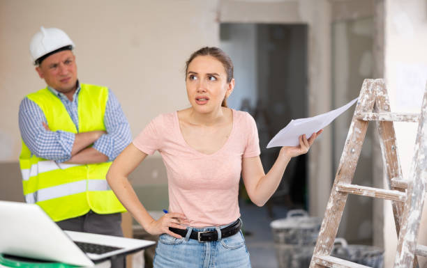 Frustrated woman holding renovation plans with a concerned contractor in a partially finished home renovation setting.